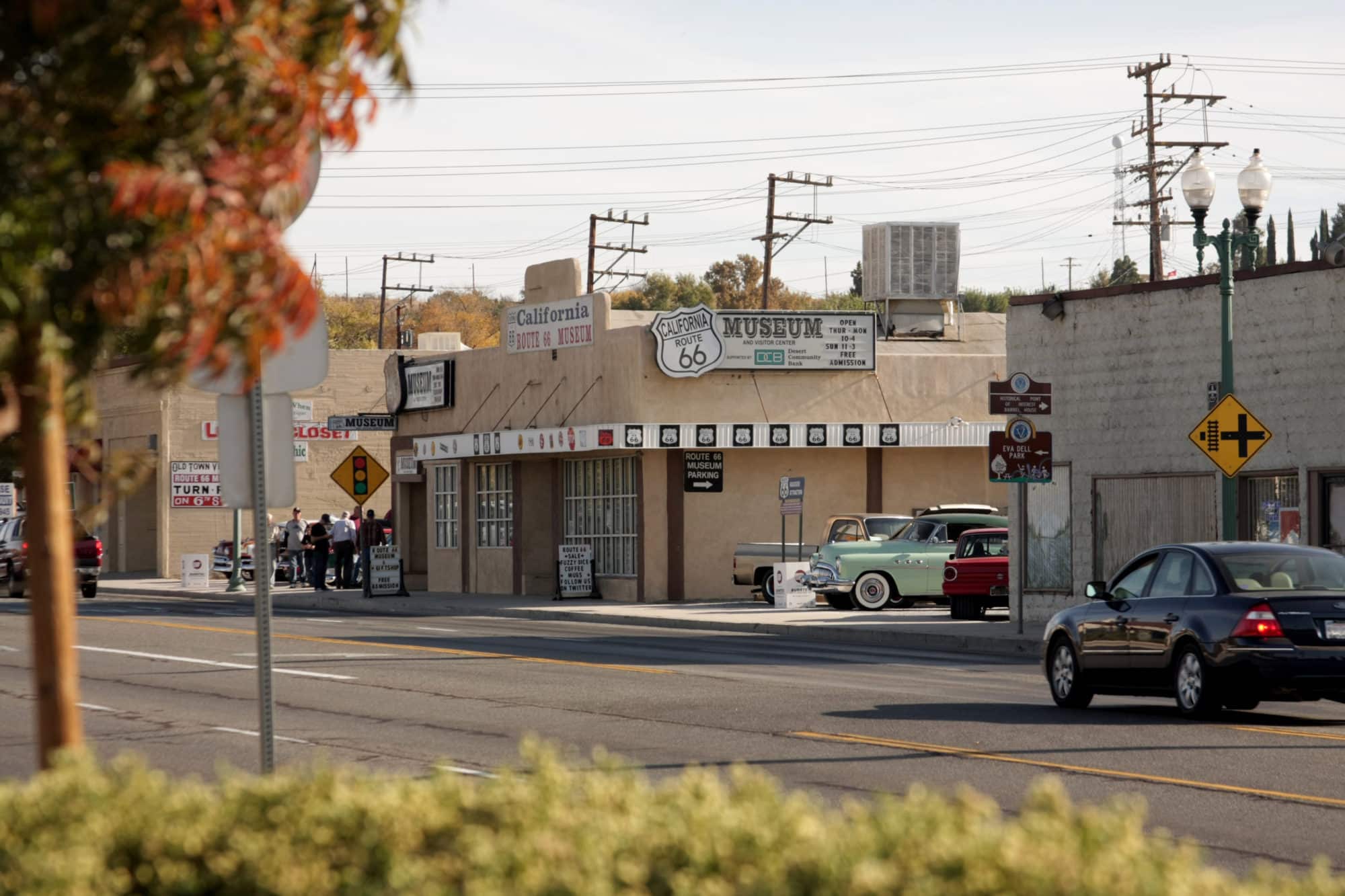 Street view of a commercial area in Victorville, California, featuring the California Route 66 Museum, vintage cars, and passing traffic.