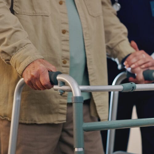 Close-up of an elderly person using a walker with support from a caregiver placing a hand on the walker for assistance.