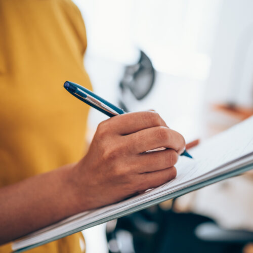 A person wearing a yellow shirt holds a clipboard and writes with a blue pen.