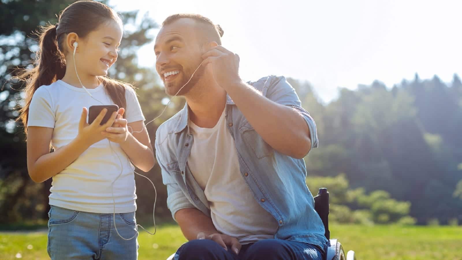A disabled father and his daughter smiling outdoors while sharing earphones and listening to music together on a sunny day.