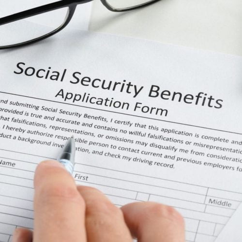 Close-up of a person filling out a Social Security benefits application form with a pen, surrounded by financial documents, charts, and a calculator.