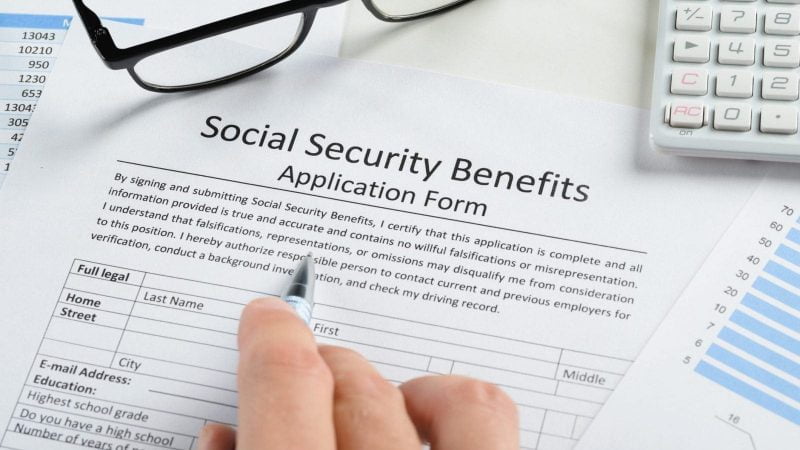 Close-up of a person filling out a Social Security benefits application form with a pen, surrounded by financial documents, charts, and a calculator.