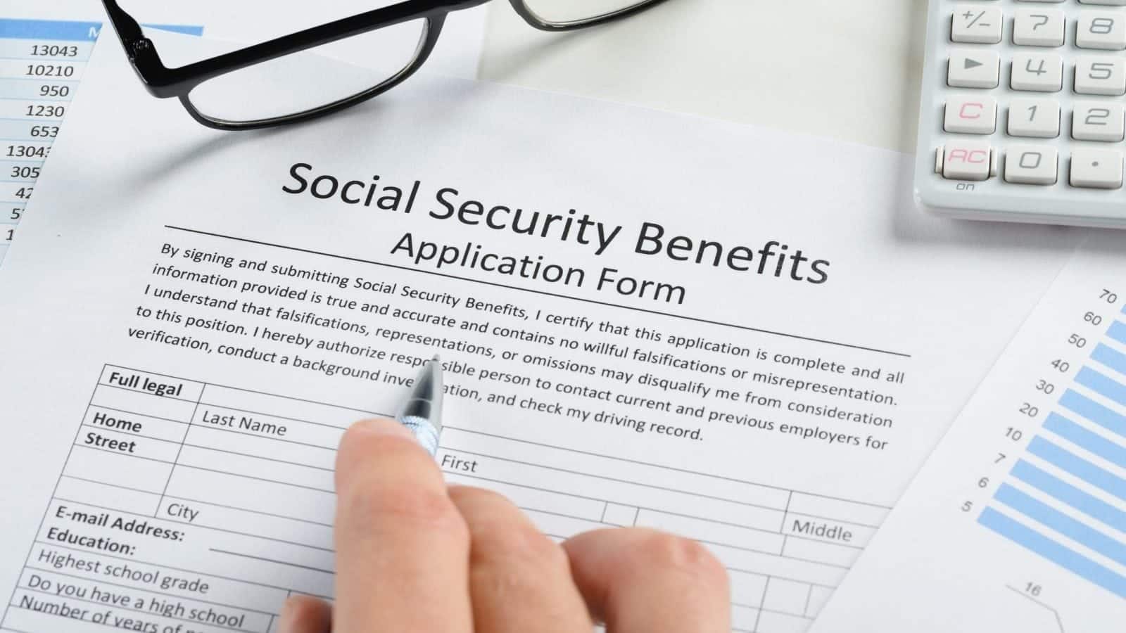 Close-up of a person filling out a Social Security benefits application form with a pen, surrounded by financial documents, charts, and a calculator.