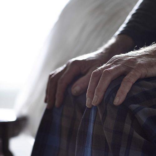 Close-up of an elderly person's hands resting on their lap while sitting on the edge of a bed.