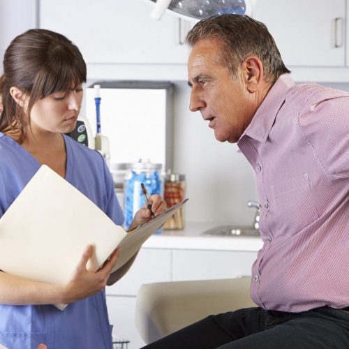 Middle-aged man holding his lower back in pain while consulting with a doctor and nurse in a medical office. The nurse is taking notes.