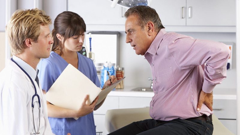Middle-aged man holding his lower back in pain while consulting with a doctor and nurse in a medical office. The nurse is taking notes.