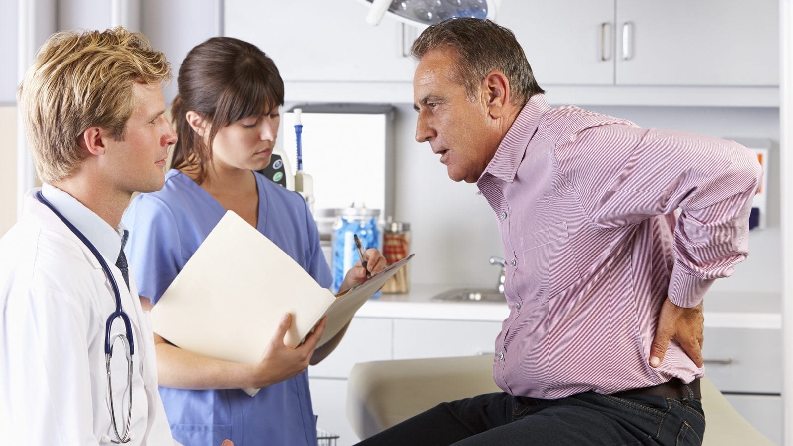 Middle-aged man holding his lower back in pain while consulting with a doctor and nurse in a medical office. The nurse is taking notes.