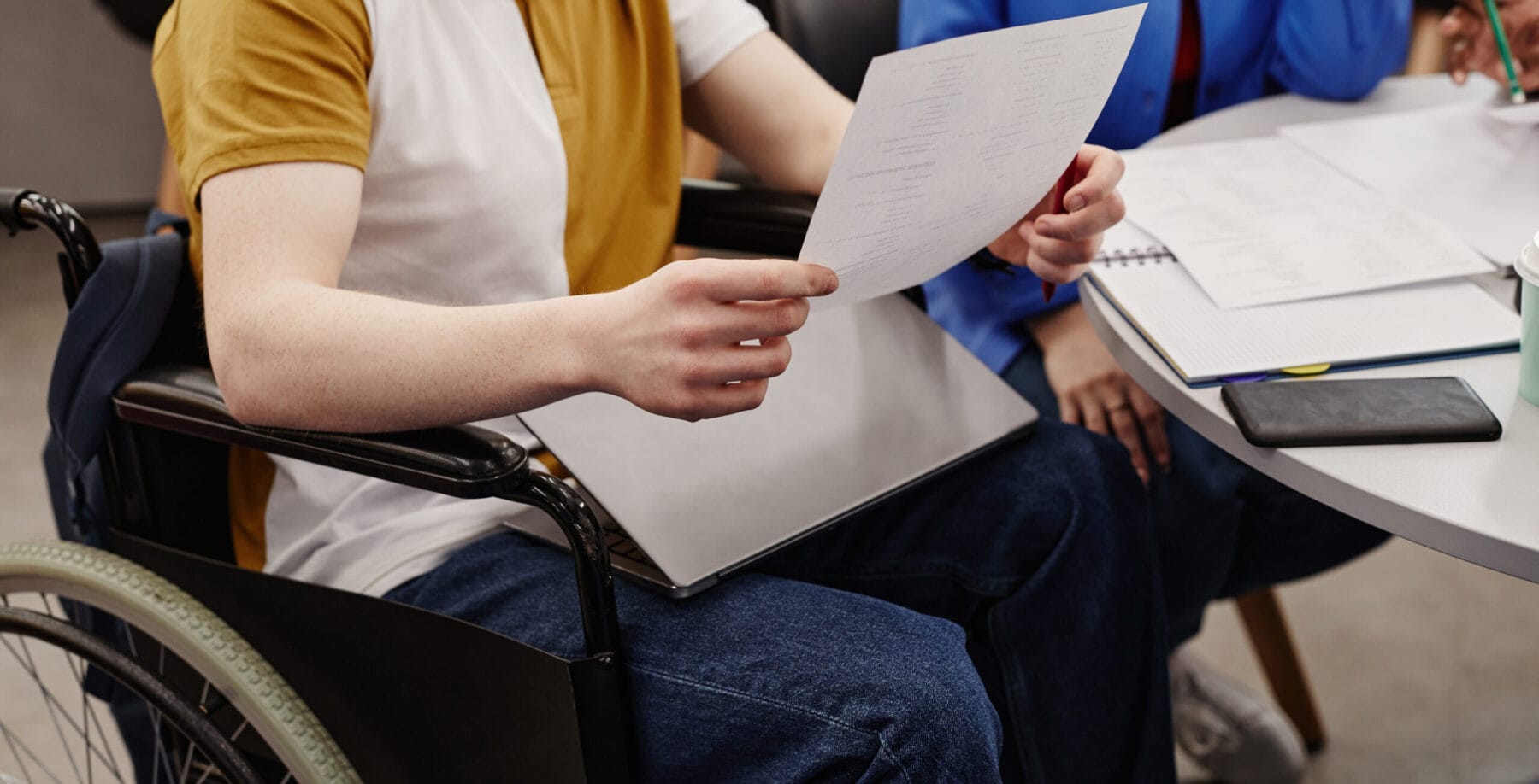 Man in a wheelchair holding paper and sitting at a desk with other people.