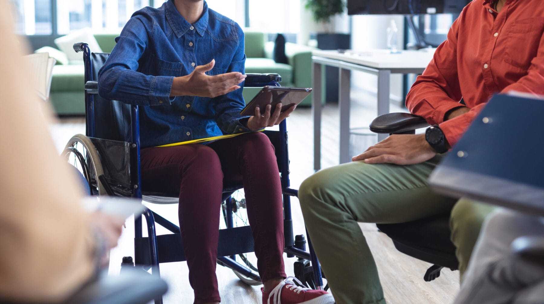 A woman in a wheelchair is holding a tablet and speaking to a group of people in an office.