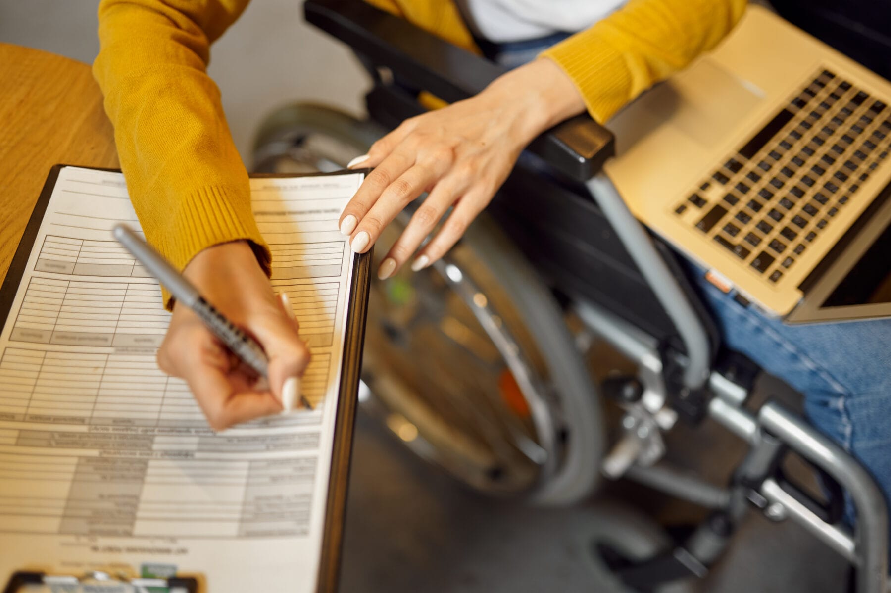 A person in a wheelchair fills out a form on a clipboard while using a laptop on their lap.