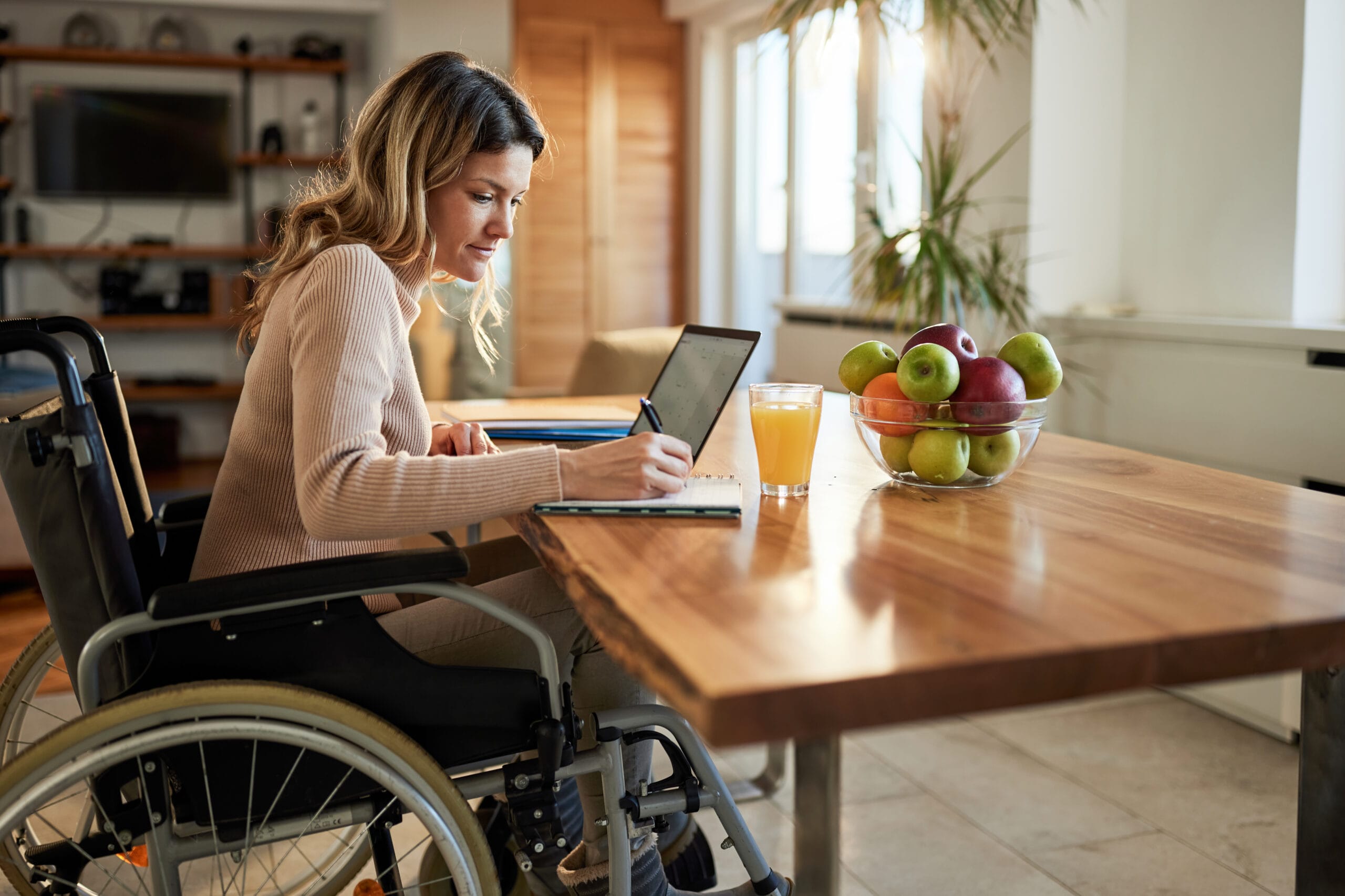 A woman in a wheelchair works at a wooden table using a laptop and notebook, with a glass of orange juice and a bowl of fruit nearby.