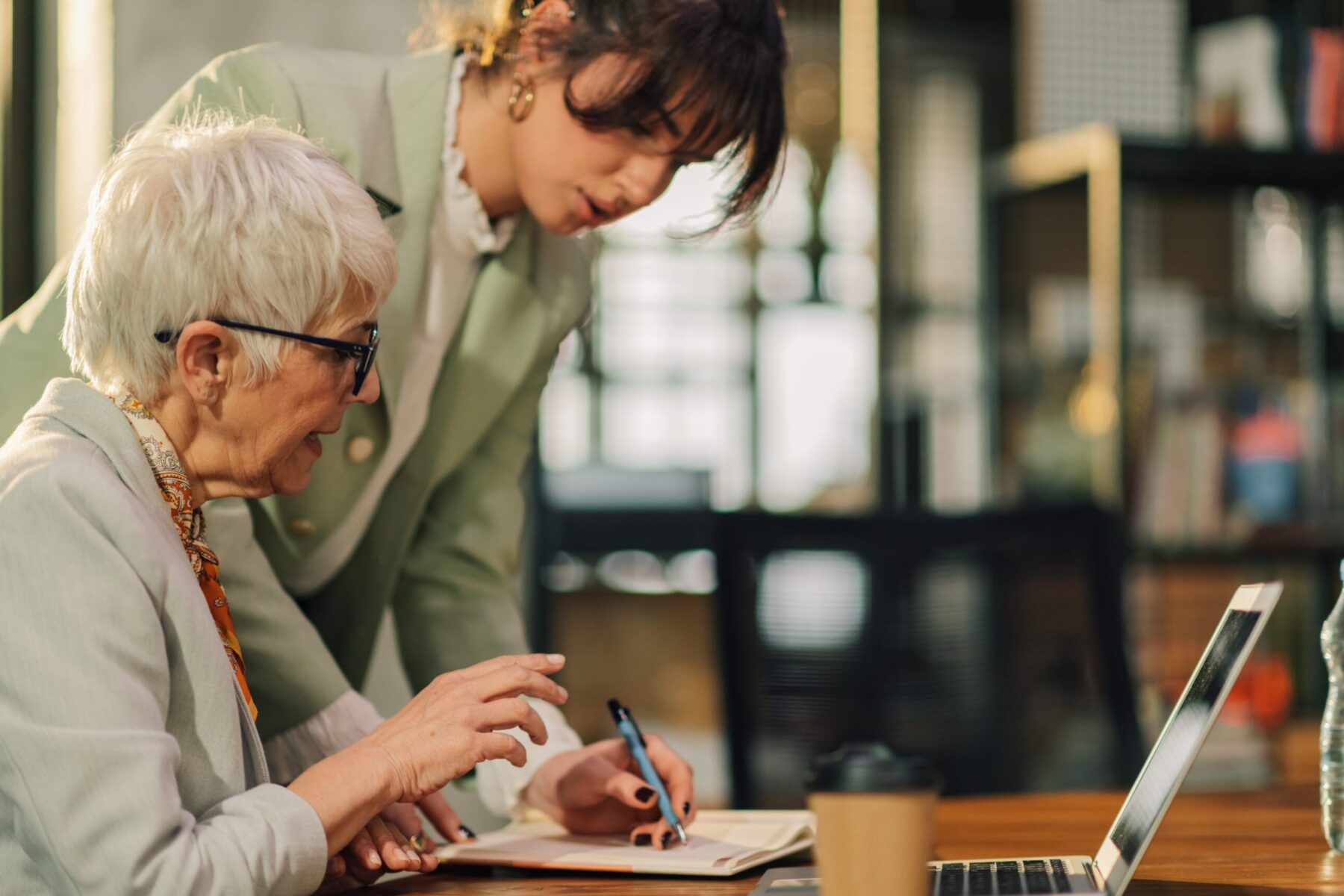 A young woman assists an older woman at a desk, reviewing notes together beside a laptop.