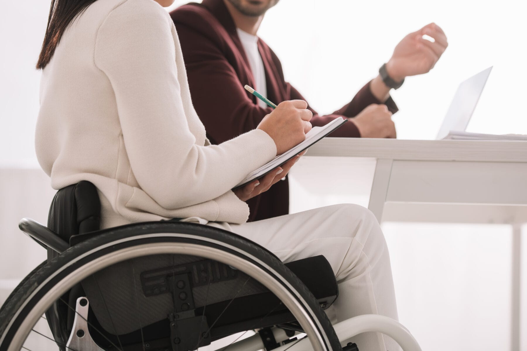 Woman in a wheelchair taking notes during a meeting with a professional at a desk.