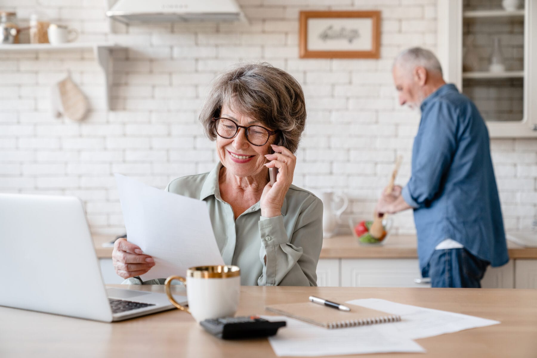 Senior woman reviewing paperwork and talking on the phone at her kitchen table while a man prepares food in the background.