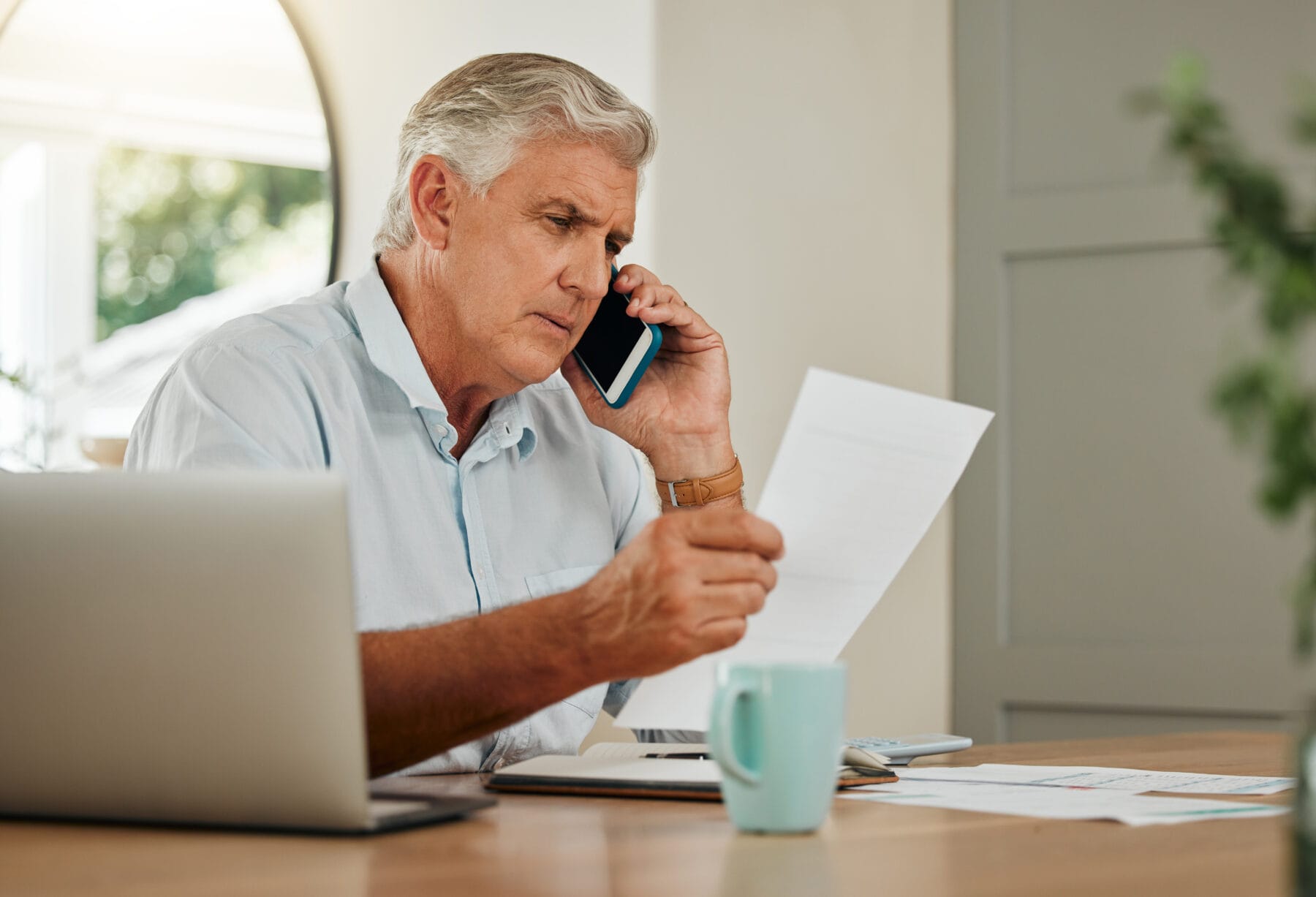 Senior man reviewing documents while talking on the phone at a table with a laptop and paperwork.