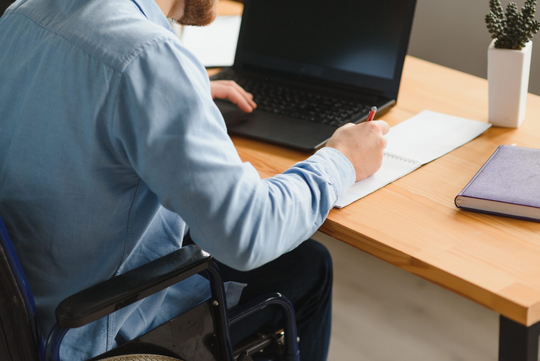 Man in a wheelchair working at a desk, writing in a notebook beside a laptop.