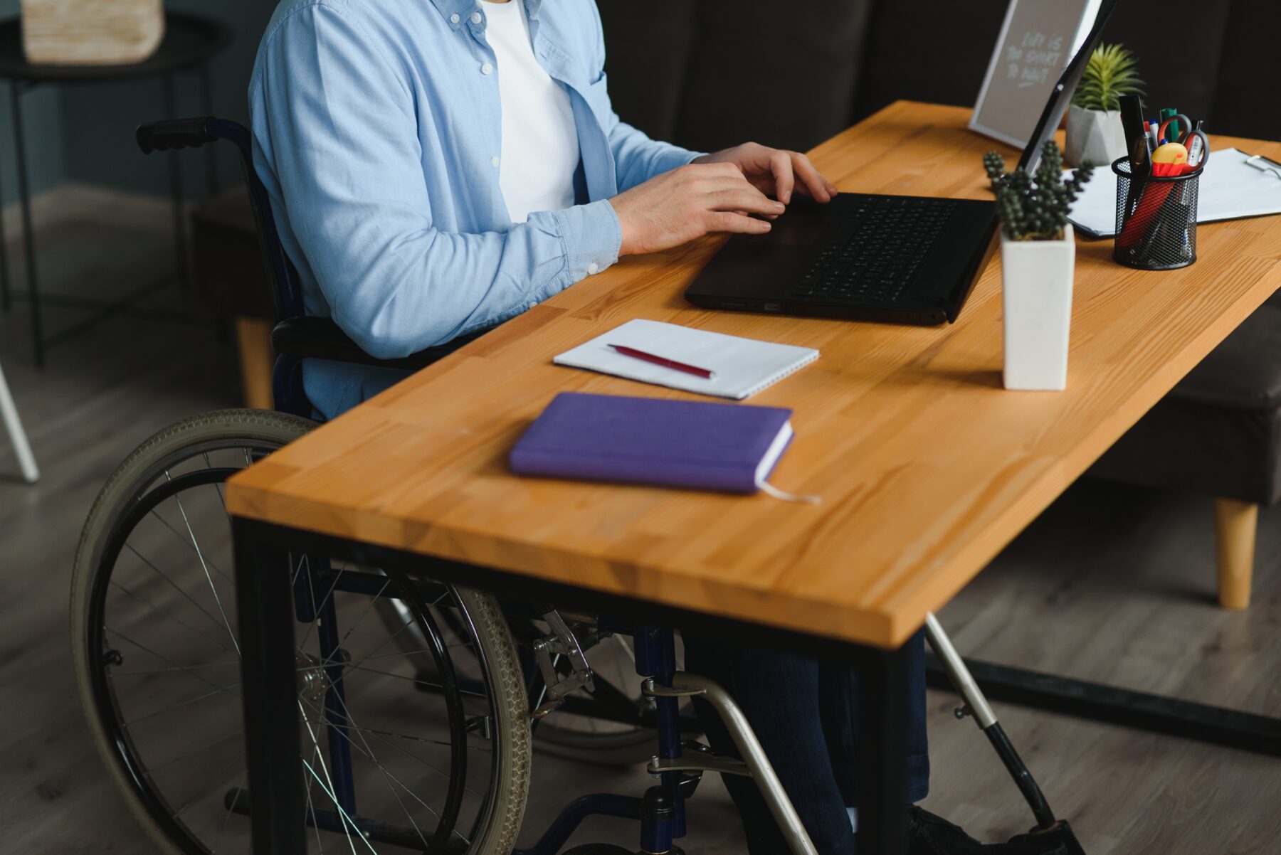 A person using a wheelchair works on a laptop at a wooden desk with a notebook and office supplies nearby.