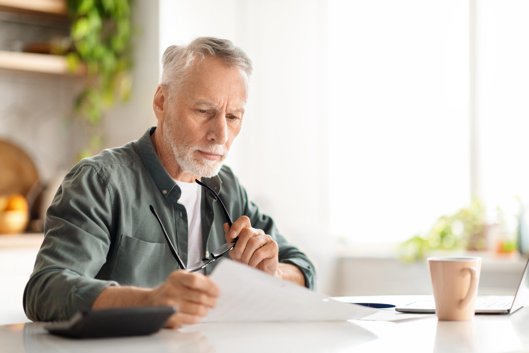 Senior man reviewing paperwork at a table while holding his glasses, with a mug and laptop nearby.